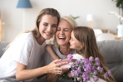 Cheerful feminine three diverse generations indoors. Sweet granddaughter mom and laughing grandmother sitting on couch in living room hold gift box and flowers embracing celebrating mother day at home