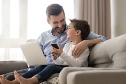 Happy young father sit on couch using laptop relax with preschooler son holding smartphone have fun together, smiling dad and little boy child enjoy weekend at home rest on sofa busy with gadgets