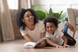 Caring african american mother baby sitter tell funny fairy tale story to smart cute kid son laugh lay on warm floor together, loving mixed race mom read book having fun with little child boy at home