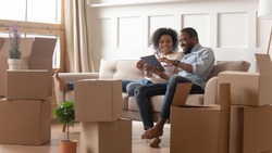 Happy young african couple use digital tablet discuss interior design renovation ideas sit on sofa on moving day, black new house renters owners tenants relax on couch with boxes having fun online