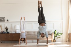 Cute active african kid girl copy imitate father doing gymnastic handstand exercise in living room, sporty family black dad and child daughter stand on hands upside down having fun together at home