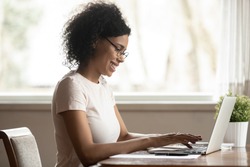 Mixed race attractive woman wearing glasses look at pc screen typing business letter informal e-mail chatting with friend online, african female freelancer sitting at table working on computer at home