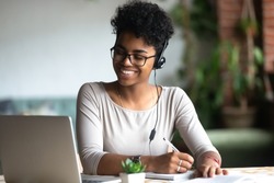 Smiling african American millennial female student in headphones and glasses sit at desk watch webinar making notes, happy biracial young woman in earphones work study using computer write in notebook