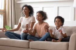 Happy peaceful young african American mom sit on couch meditate together with little children, smiling calm black mother or nanny relax practice yoga with mudra hands teach small boy and girl kids