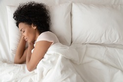 Serene calm african american woman sleeping in comfortable bed lying on soft pillow orthopedic mattress, peaceful young black lady resting covered with blanket on white sheets in bedroom, top view