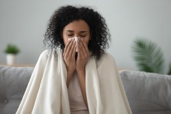 Ill african young woman covered with blanket blowing running nose got fever caught cold sneezing in tissue sit on sofa, sick allergic black girl having allergy symptoms coughing at home, flu concept