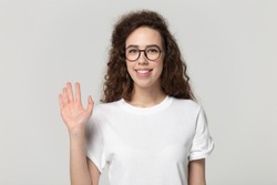 Smiling 20s girl wearing glasses white t-shirt posing isolated on gray studio background, woman looking at camera wave hand greeting get acquainted with someone polite gesture nice to meet you concept