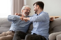 Happy old senior father and son fists bumping, celebrating success or greeting each other, mature aged dad and millennial man having fun together, sitting on couch at home, enjoying weekend