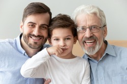 Head shot portrait smiling grandfather, father and preschool son looking at camera, bearded granddad, dad and little grandchild boy with painted fake mustache on finger posing for funny family photo