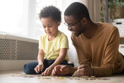 Loving african American dad lying on floor with biracial little son engaged in game assemble jigsaw together, happy black father play have fun with preschooler kid make picture connect puzzle pieces