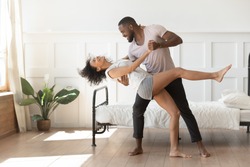 Cheerful active romantic african american couple wearing pajamas dancing in bedroom together, happy carefree young black husband and wife enjoy weekend morning laughing bonding having fun at home