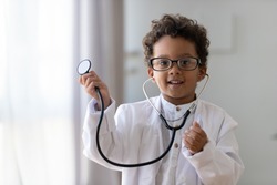 Cute small african american kid boy wear medical uniform glasses holding stethoscope playing doctor, happy funny little mixed race preschool child pretending pediatrician looking at camera, portrait