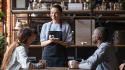Smiling millennial waitress with notebook taking order from young multiracial client couple, diverse friends relax hang out in cafe or restaurant speak with staff enjoying good service and atmosphere