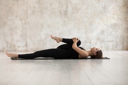 Woman wear black sport clothes lying on floor practising asana do Half Knees to Chest Pose near grunge wall beige textured background, help ease back pain, flexible body stretch for beginners concept