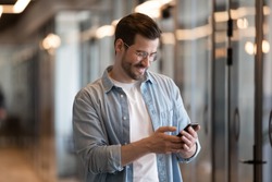 Happy young businessman using smart phone standing in office hallway, smiling male professional holding smartphone working with business apps texting sms enjoy corporate technology on mobile device
