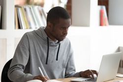 Serious concentrated african american male student using laptop, busy with studying, preparing for evaluation examination, final test or session. Motivated black guy doing homework or writing essay.