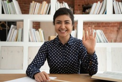 Happy indian female student looking at camera, waving hello, greeting. Smiling cheerful young intern holding interview work call with potential employer, recording self-introduction video in library.