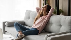Serene young woman pretty relaxing on couch in living room. Calm female freelancer crossing hands behind head, closing eyes and taking a break during remote work day at home, stress free concept.