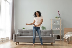 Overjoyed black biracial millennial girl dancing alone moving to rhythm in living room, happy african American young woman have fun enjoying listening to music, entertain on weekend at home