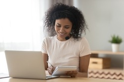 Happy african American female student wearing headset writing down in notebook busy studying on laptop, smiling black woman take online learning course make notes, watch webinar on computer