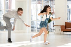 Cute child daughter running from happy dad catching playing tag and touch game at home, father chasing excited kid girl having fun enjoy leisure activity laughing spend time together in living room