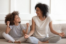 Excited african american woman teaching cute little adorable daughter meditating and practicing yoga exercises. Happy mixed race family sitting on comfy couch in lotus position, having fun together.