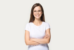Smiling beautiful millennial girl wear glasses and white t-shirt stand with arms crossed isolated on grey studio background, happy young woman in spectacles look at camera posing or casting