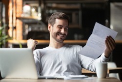 Excited business man student reading postal mail letter overjoyed by great news, happy male winner holding paper bill with loan approval celebrate taxes refund receive salary rise payment sit at desk