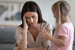 Annoyed mom and restless little daughter at home, mother sitting on couch touch head or temples feels headache due to noisy kid standing near her, tired parent of difficult hyperactive child concept