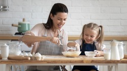 Pretty woman and little adorable daughter sitting at table in kitchen cook together stirring eggs for pie, cookies or pancakes, teach kid recipes, embodiment of ideal wife of mom and housewife concept