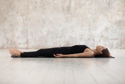 Young woman wearing black sportswear practicing yoga, doing Corpse, Savasana exercise, relaxing, lying in Dead Body pose on mat, sporty girl working out at home or in yoga studio with grey walls