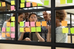 Focused multiethnic business team people brainstorm on corporate project together write strategy tasks on post in sticky notes stand behind glass wall, diverse staff group planning management concept