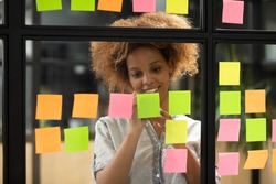 Smiling young african american businesswoman student teacher coach writing creative ideas on sticky post it notes on glass scrum board wall working on project strategy plan in modern corporate office