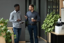 Diverse friendly male partners colleagues talking walking in modern office hallway, young african american and caucasian business men discussing common project work meeting in company work space