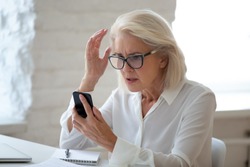 Confused senior businesswoman sit at office desk hold cellphone experience internet connection problem, frustrated aged woman worker feel disappointed having smartphone breakdown or virus attack