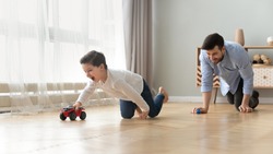 Funny happy male family young adult dad and cute excited little kid son pretending racing on warm wooden floor at home, father having fun chasing small preschool child boy playing cars together