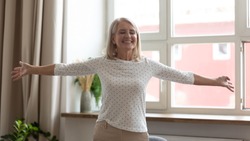Happy smiling mature woman standing with arms outstretched at home, thankful satisfied older female with closed eyes enjoying life, having fun, rejoicing success, weekend or retirement