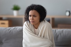 African unhappy woman sit on couch covered with plaid try to warming up in cold flat without central heating, system not working, girl feels unhealthy has common cold viral infectious disease concept