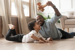 Happy smiling loving black single father in eyeglasses holding wooden plane in air, playing with little mixed race cute curly funny child son, lying on heated floor, spending weekend leisure time.