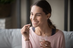 Smiling healthy young woman holding pill glass of water sit on sofa at home, positive lady take daily medicine antioxidant diet vitamin supplements for beauty skin hair health care medicament concept