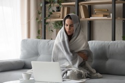 Sick ill young woman feel cold covered with blanket sit on sofa watching movie on laptop, annoyed girl shiver freezing warming at home wrapped with plaid, no central heating problem and flu concept