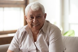 Elderly positive happy man seated on couch in living room smiles looking at camera feels healthy, 80s grandfather having wide toothy smile, concept of dental care exam for senior clinic advertisement