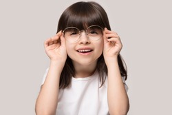 Headshot portrait of cute little preschooler girl isolated on grey studio background wear glasses look at camera, small child try spectacles at opticians, kid eyesight correction treatment concept