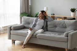 Full length positive dreamy young biracial girl leaning on comfortable couch, holding mobile phone in hands, looking at window, thinking of future date, meeting with friends or enjoying calm moment.