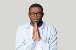 Head shot portrait upset African American man in glasses joining hands in prayer gesture with hope isolated on grey studio background, unhappy young male with closed eyes asking help and support