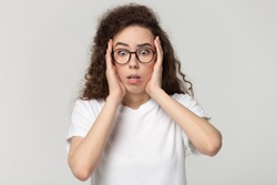 Frustrated teenage girl wearing glasses isolated on grey studio background touch head feel terrified or scared, shocked millennial woman in spectacles fence herself frightened or paralyzed with idea