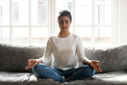 Full length mindful young indian woman making mudra gesture, sitting in lotus position on comfortable couch at home. Peaceful millennial girl deeply meditating, doing breathing yoga exercises alone.
