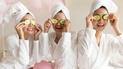 Happy young women wear white bathrobes towels on head make cucumber facial skin care mask on eyes laughing relaxing together, smiling girls friends having fun on spa beauty salon party with balloons