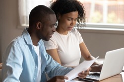 Multiracial young couple sit at table at home managing paperwork financial documents consider family expenditures together, multiethnic husband and wife pay household taxes or bills on laptop online