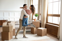 Happy young husband lifting excited wife celebrating moving day with cardboard boxes, proud overjoyed family couple first time home buyers renters owners having fun enjoy relocation, mortgage concept